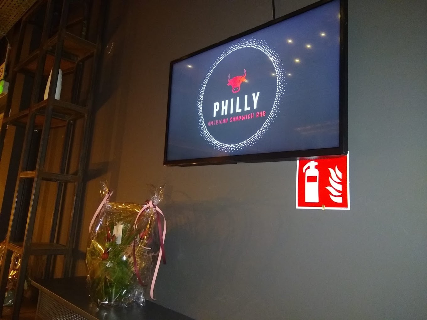 The interior of halal restaurant Philly Sandwich Bar in Antwerpen, Belgium, showing a television screen with the restaurant's logo and a fire safety sign on the wall, along with a floral arrangement on a table.