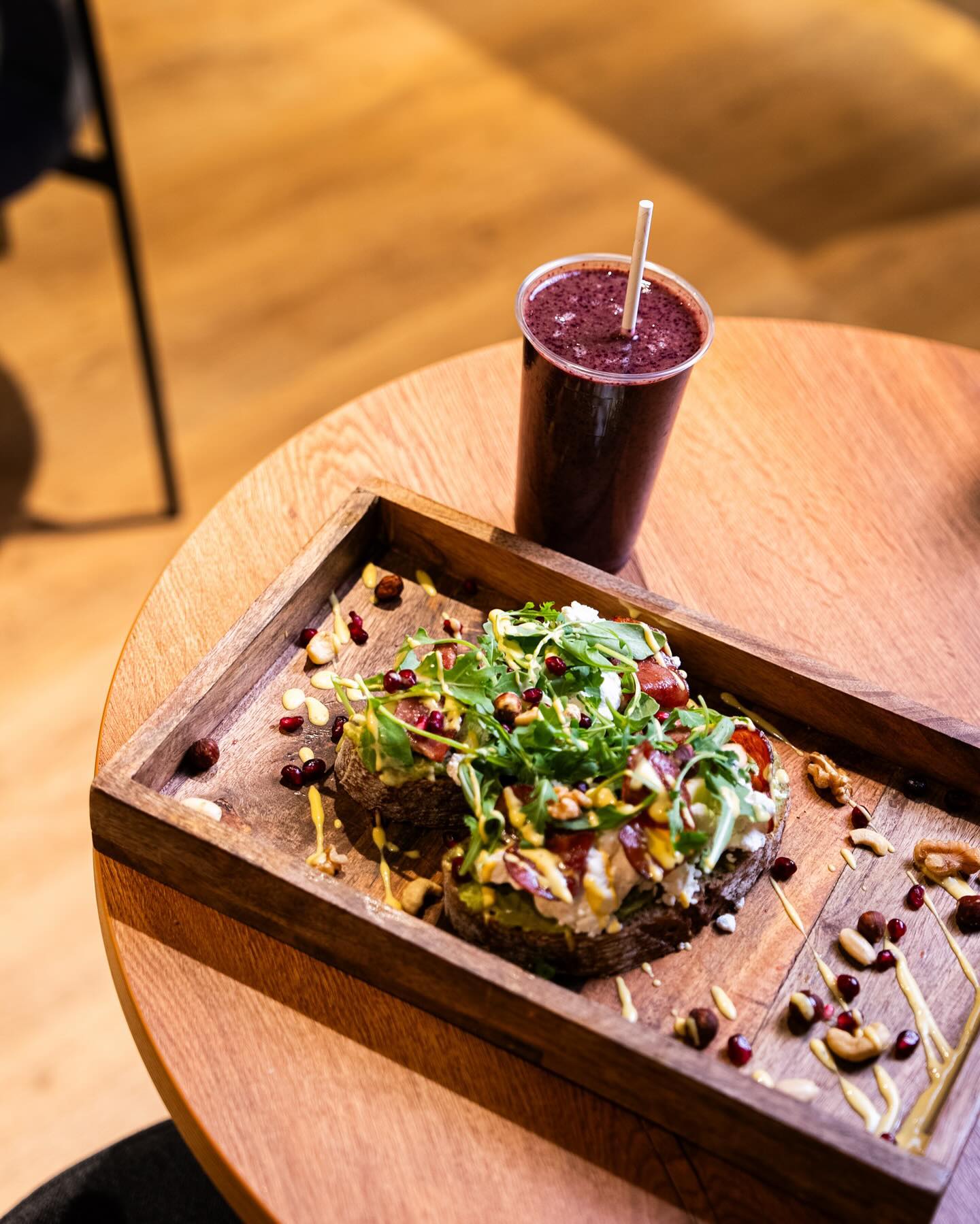 A wooden tray with two slices of dark bread topped with greens, pomegranate seeds, and a drizzle of sauce, accompanied by a smoothie at Freshmade, a halal restaurant in Wijnegem, Belgium.