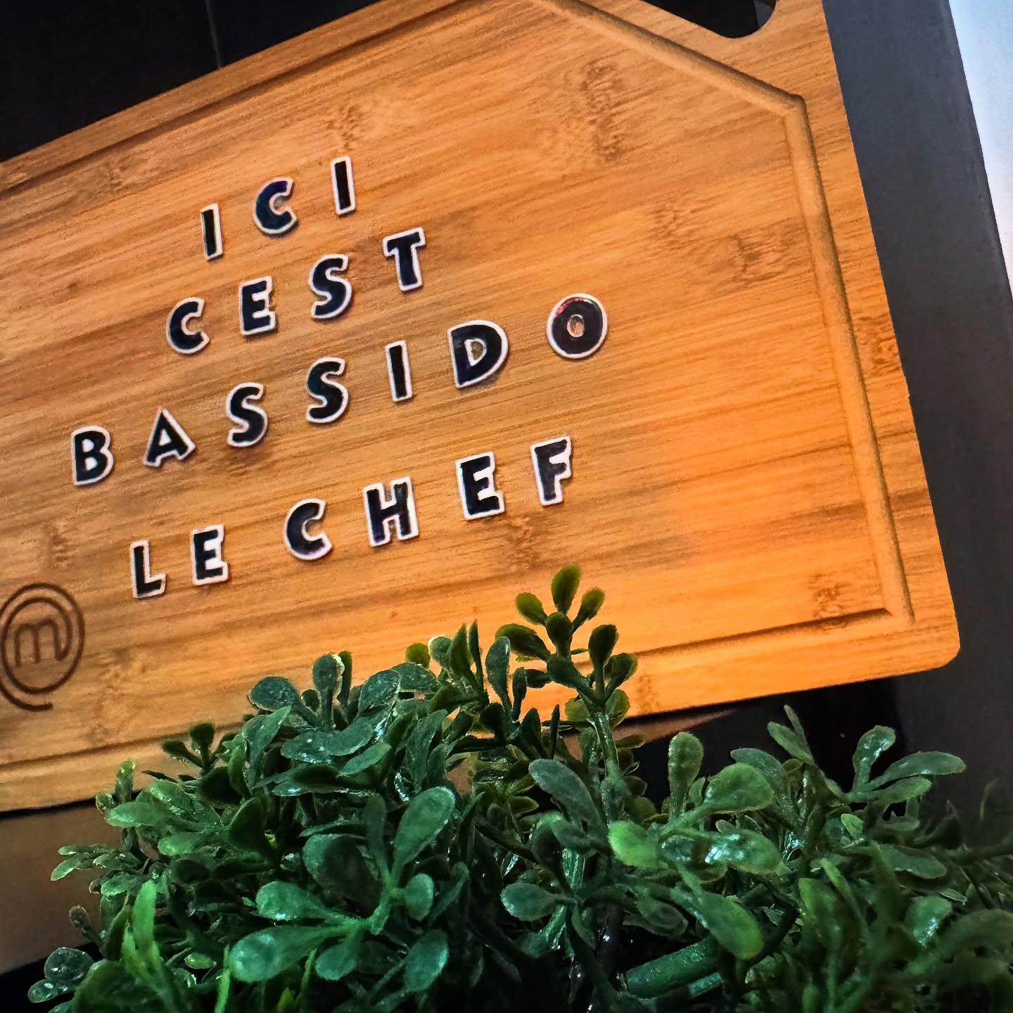 The interior of halal restaurant Baba Brunch in Koekelberg, Belgium, showing a wooden sign with the text 'ICI C'EST BASSIDO LE CHEF' and a small green plant in the foreground.