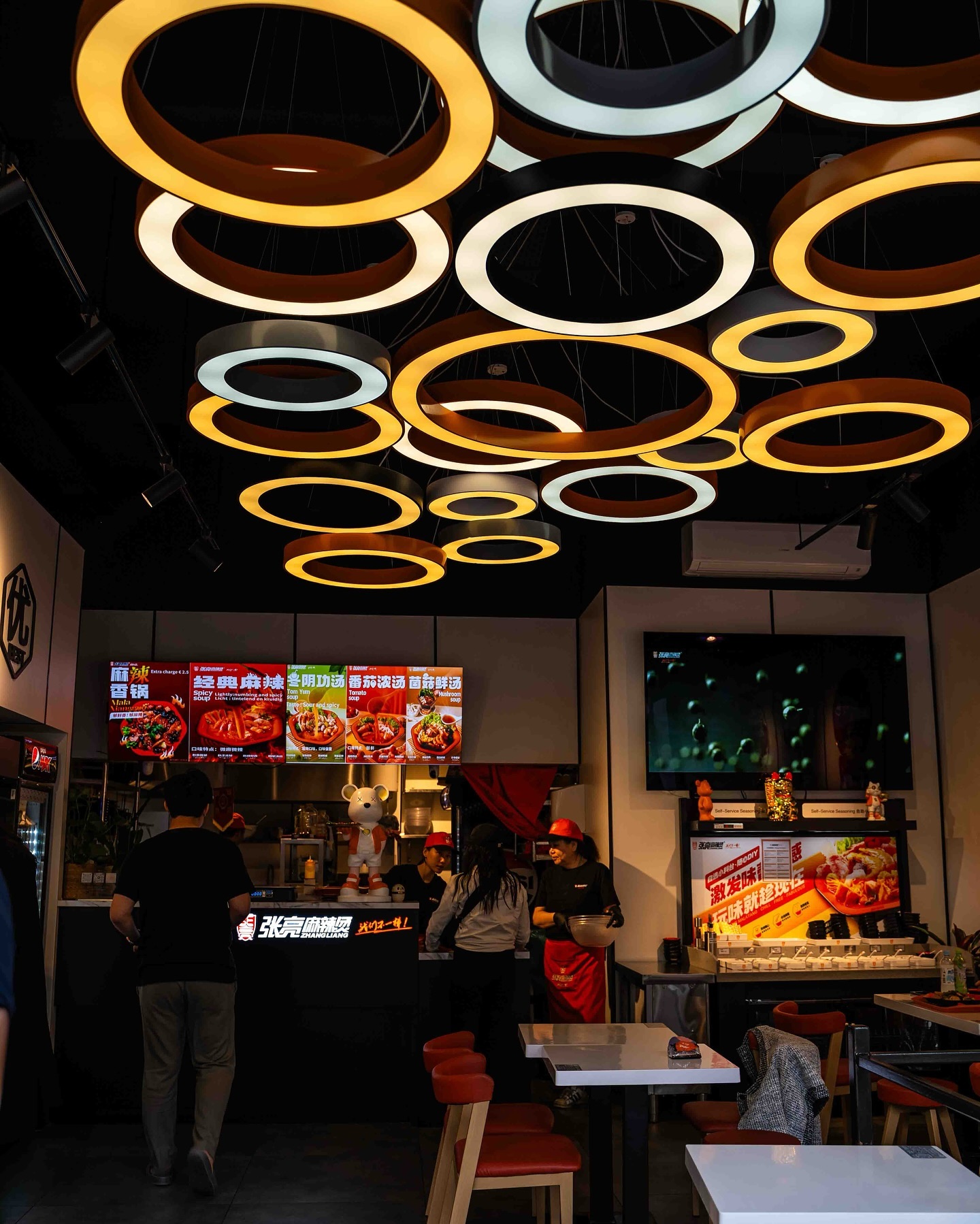 The interior of halal restaurant ZLMLT in Rotterdam, Netherlands, showing a modern design with circular light fixtures, a counter with menu displays, and staff preparing food.