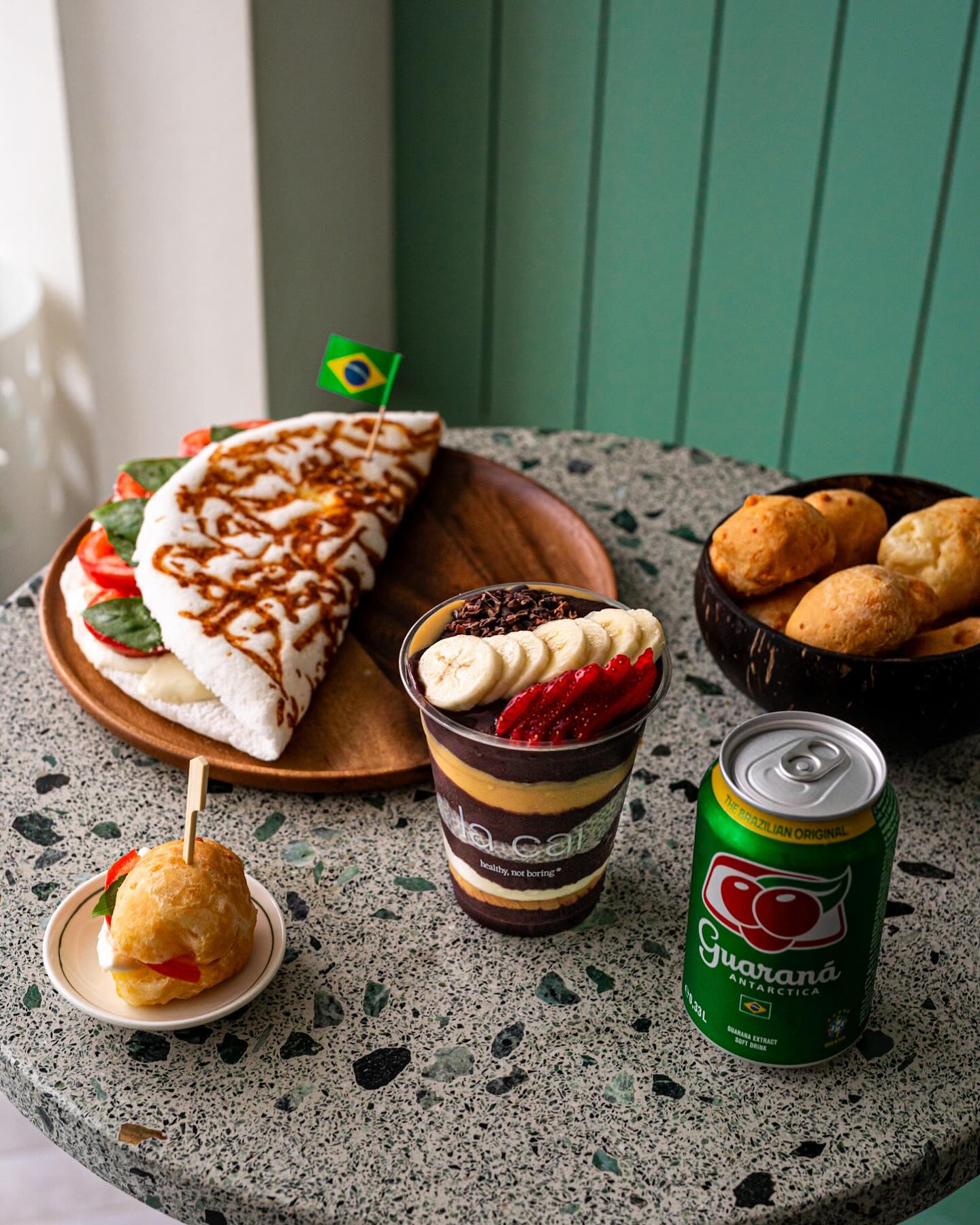 Une assiette de nourriture brésilienne comprenant une tapioca farcie, un verre de dessert avec des couches de chocolat et de fruits, une petite pâtisserie et une canette de Guaraná au restaurant halal La Cai à Bruxelles, Belgique.