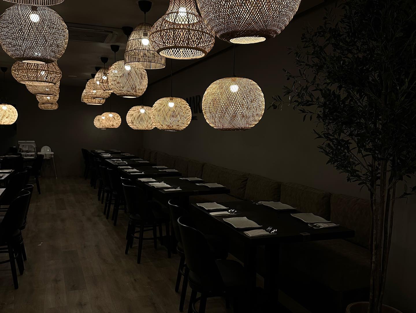 The interior of halal restaurant Al Mar in Antwerpen, Belgium, showing a row of tables with black chairs, a green seating area, and decorative woven pendant lights hanging from the ceiling.