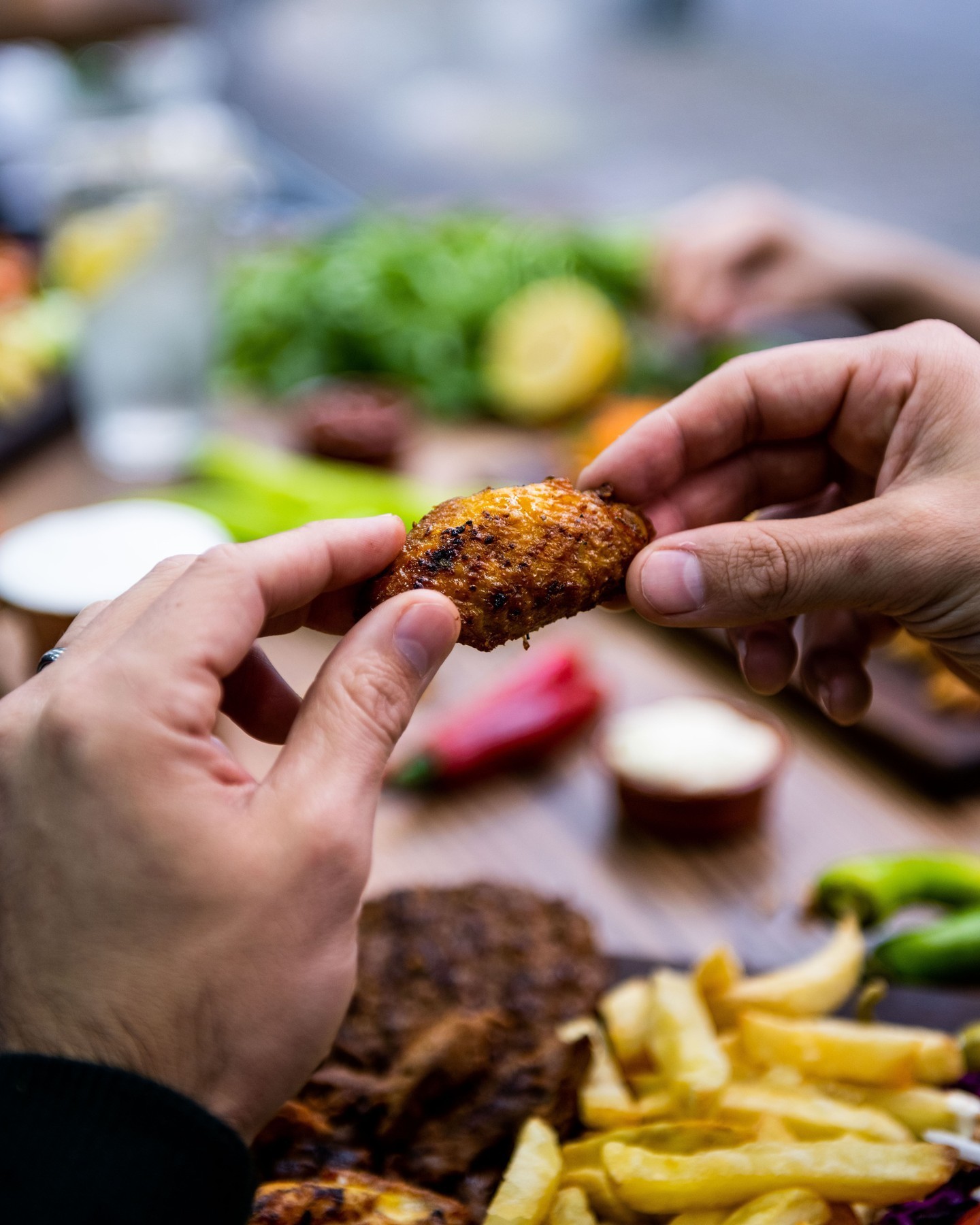 Un gros plan d'une main tenant une aile de poulet croustillante, avec une variété de plats d'accompagnement visibles en arrière-plan au Snack Murat, un restaurant halal à Saint-Gilles, Belgique.