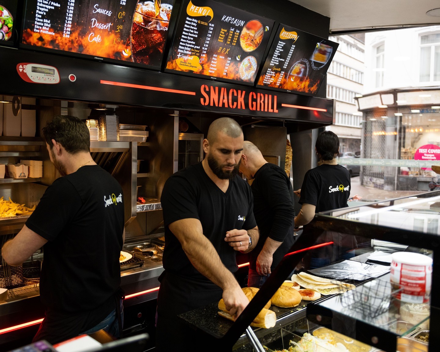 L'intérieur du restaurant halal Snack Murat à Saint-Gilles, en Belgique, montrant le personnel préparant des plats derrière un comptoir avec un menu affiché sur des écrans au-dessus.