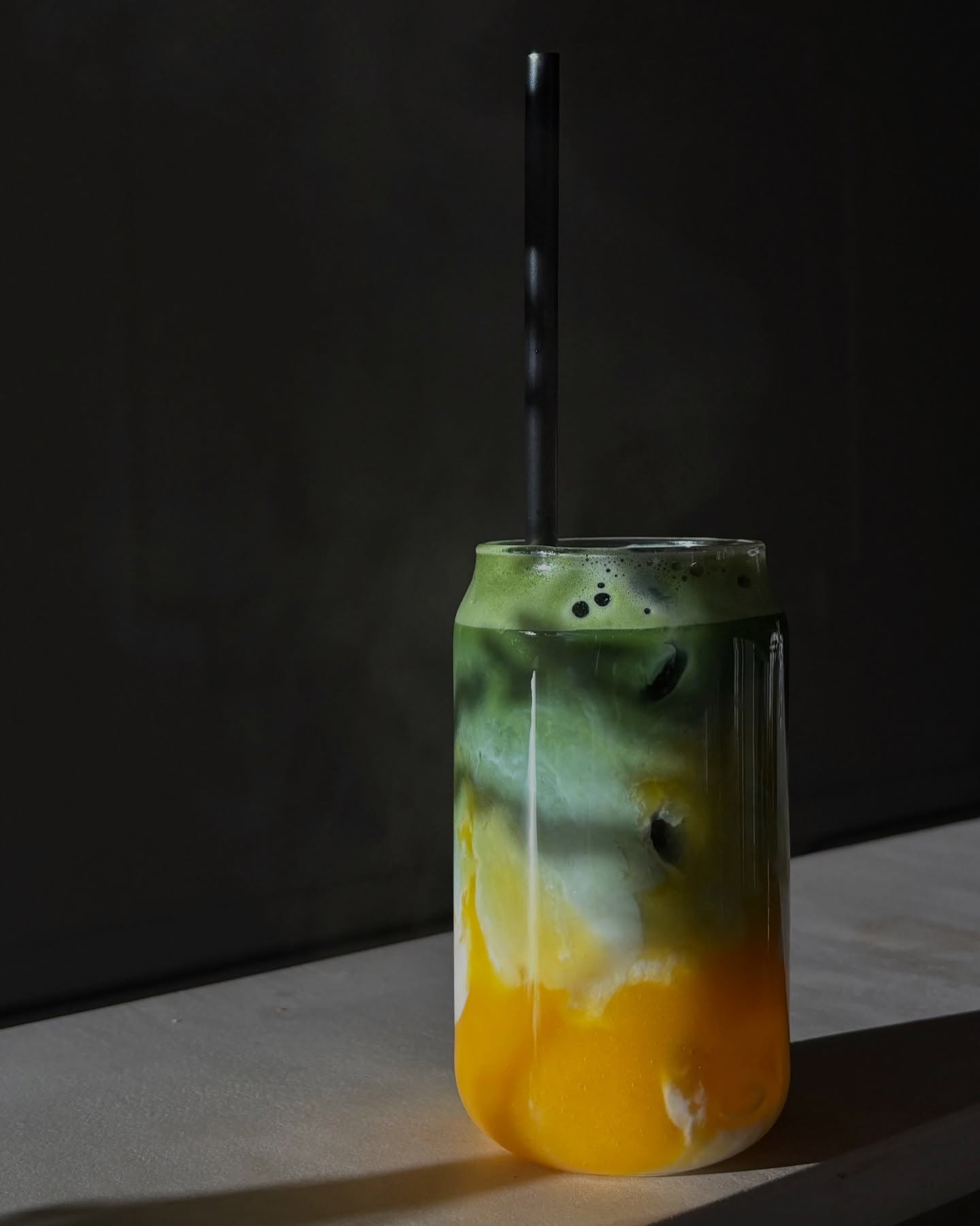 Une boisson en couches avec des teintes vertes et orange, surmontée d'une mousse, servie dans un verre avec une paille noire au restaurant halal Mio à Anvers, Belgique.