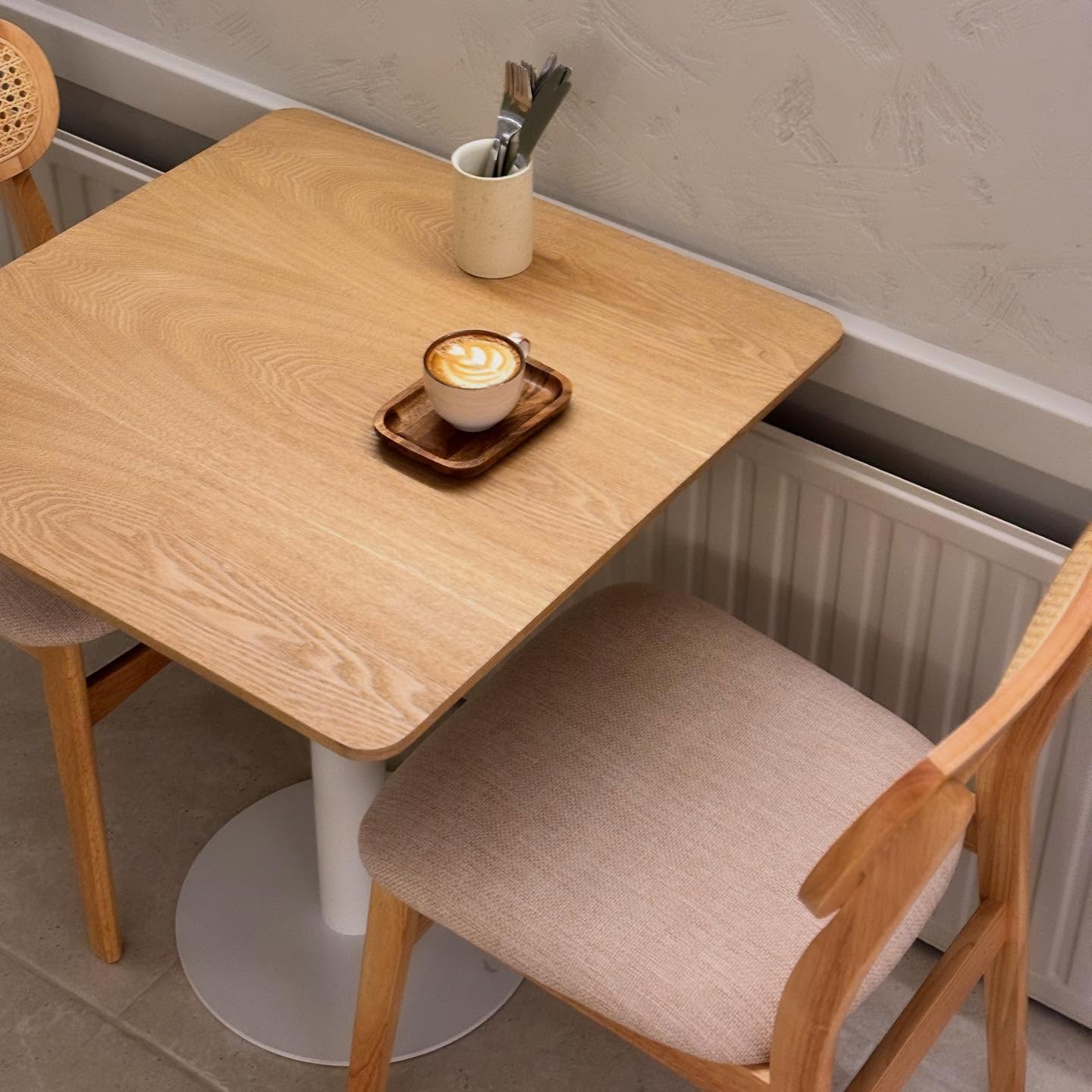L'intérieur d'un café montrant une table en bois avec une tasse de café sur un plateau, entourée de deux chaises en bois et d'un porte-ustensiles.